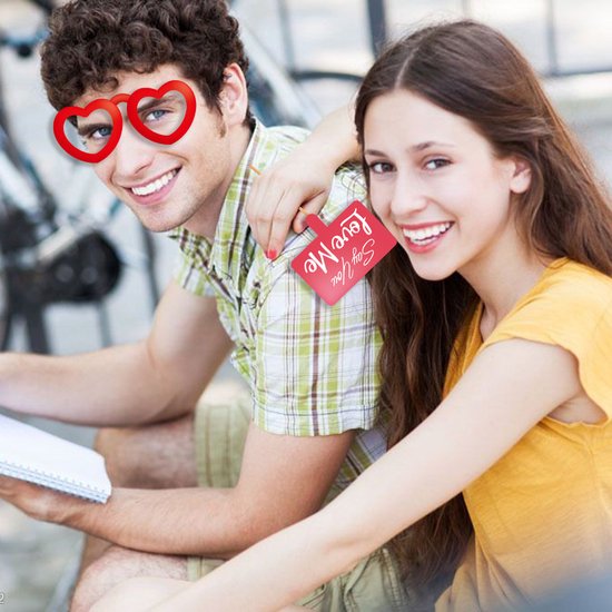Een man en vrouw zitten buiten, de man draagt een hartvormige bril en de vrouw houdt een rood kaartje met 'Say You Love Me' vast.