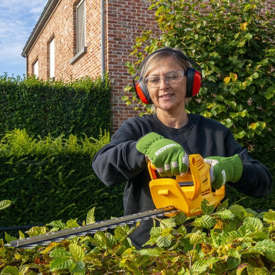 Vrouw met gehoorbescherming en veiligheidsbril gebruikt gele heggenschaar bij een tuinhek.