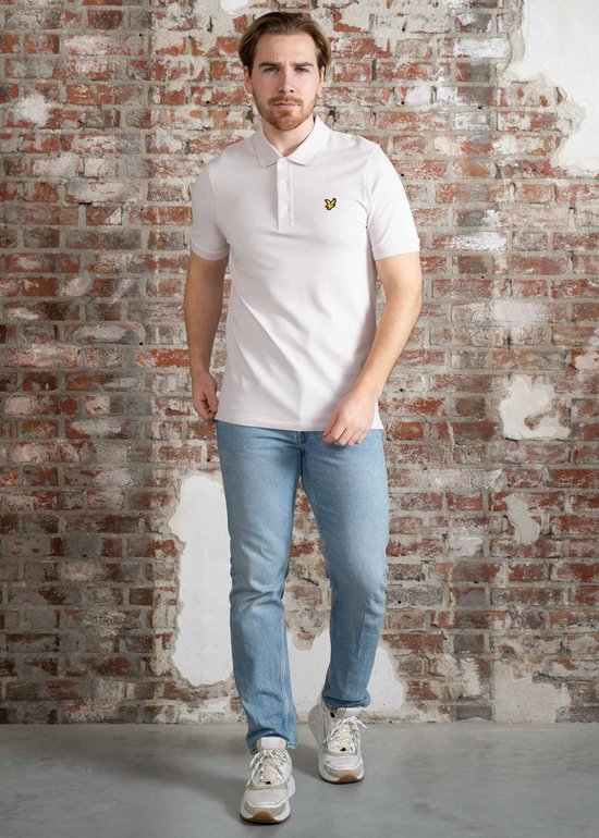 Man in lichtpaars Lyle & Scott poloshirt met korte mouwen en gele vogel-logo op de borst, staand voor een bakstenen muur.