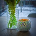 Een ronde mozaïek waxinelichthouder met een groen-oranje gloed naast een glazen vaas met groene planten.