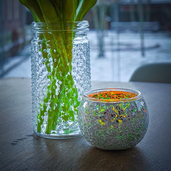 Een ronde mozaïek waxinelichthouder met een groen-oranje gloed naast een glazen vaas met groene planten.