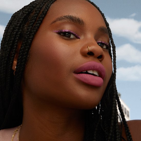 Model met lange vlechten en paarse eyeliner, draagt zachtroze lippenstift.