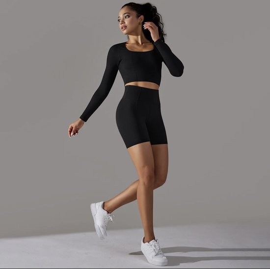 Model in een zwarte longsleeve crop top en zwarte biker shorts, gedragen met witte sneakers.