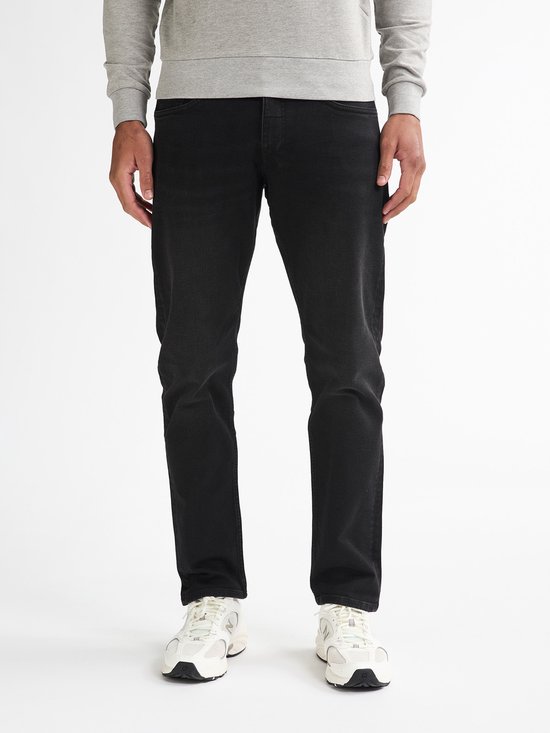 Man van voren gezien, draagt een grijze sweater en zwarte jeans met witte sneakers.
