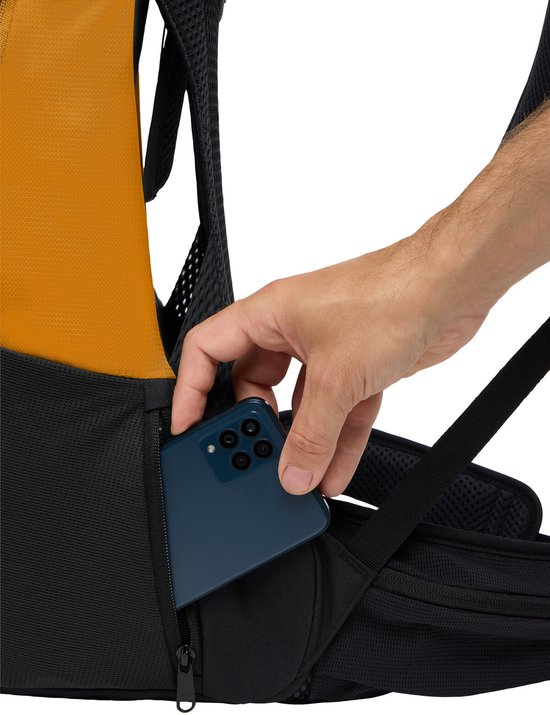 Hand doet smartphone in een ritsvak van een oranje rugzak met gaasdetails.