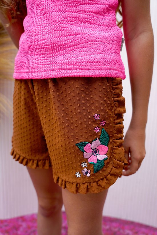 Bruine shorts met stipjes, ruches en geborduurde bloemen op de linkerpijp. Een roze top met structuur is zichtbaar.