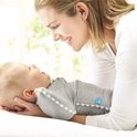 Vrolijke baby ligt in grijze Swaddle UP slaapzak bij een lachende vrouw, in het zachte licht bij een raam.
