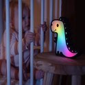 Een veelkleurig, oplichtend dinosaurusspeeltje staat op een houten nachtkastje naast een baby in een ledikant.