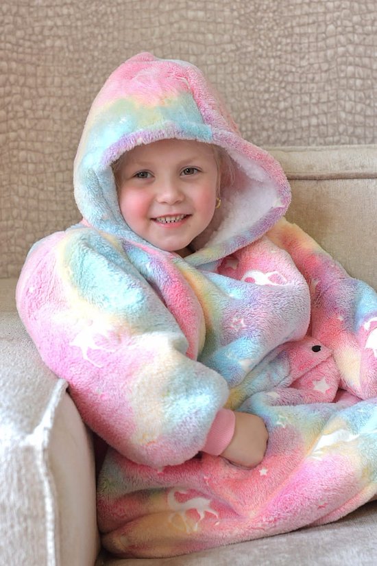 Close-up van een jong meisje met blonde staartjes, zittend op een bank, in een zachte pluche hoodie deken met pastel regenboogkleuren, sterren en eenhoorns.