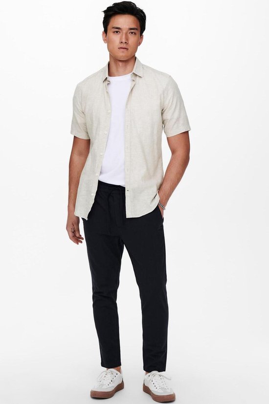 Man met een beige linnen mix overhemd met korte mouwen, een witte t-shirt en een zwarte broek.