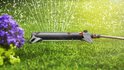 Grijze zwenksproeier sproeit water op groen gras, met paarse bloemen op de voorgrond.