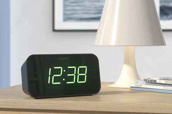 Zwarte Philips wekkerradio met groene digitale tijd op een houten nachtkastje, naast een lamp en boeken.