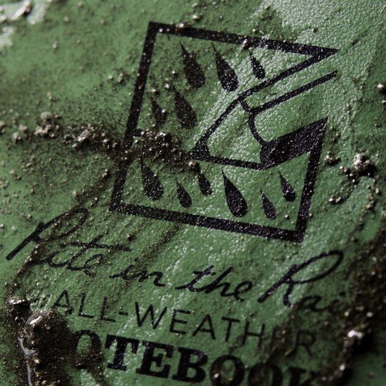 Close-up van het Rite in the Rain logo op een groen, waterdicht notitieboek, met kleine metaaldeeltjes erop.