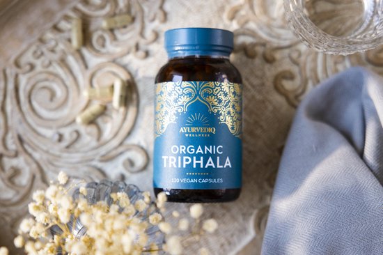 Bruine fles met blauw, goudkleurig etiket met Ayurvediq Organic Triphala, 120 vegan capsules, op een sierlijke ondergrond.