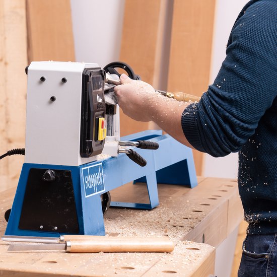 Persoon in blauwe trui die hout bewerkt op een blauwe Scheppach houtdraaibank. Veel houtsnippers op de werkbank.