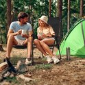Man en vrouw zitten in zwarte campingstoelen in bos, met tent en kampvuur.
