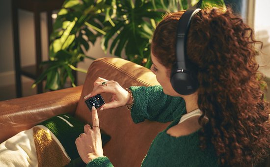 Vrouw met krullend rood haar luistert naar muziek op JBL koptelefoon, terwijl ze een afspeelapparaat vasthoudt.