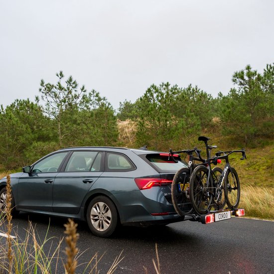 Twee zwarte racefietsen op een trekhaakfietsendrager achter een donkergrijze stationwagen, met duinen en pijnbomen op de achtergrond.