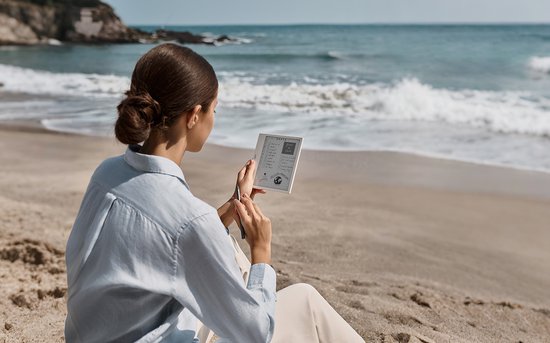 Vrouw met bruin haar in een blauwe blouse op het strand, leest op een witte e-reader.