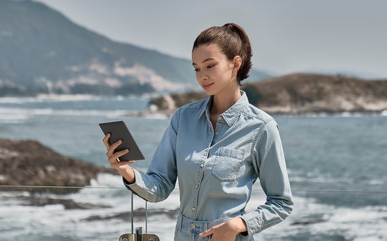 Vrouw met paardenstaart in spijkerblouse leest op een donkere e-reader aan zee.