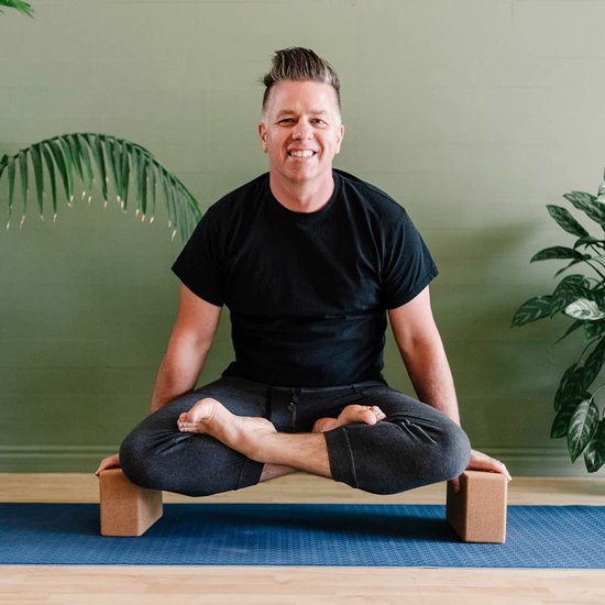 Homme aux cheveux coiffés en pics, en t-shirt noir et pantalon gris, assis en tailleur sur un tapis de yoga bleu avec des blocs marron.