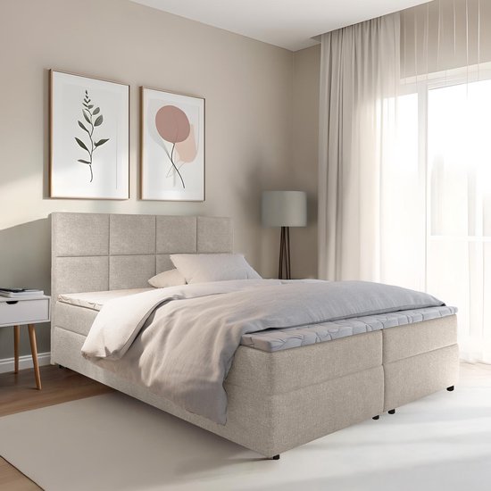 Opbergboxspring van lichtbeige stof met een gestoffeerd hoofdbord. Het bed is opgemaakt met beddengoed.