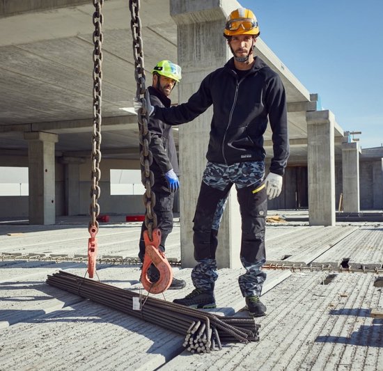 Twee bouwvakkers op een bouwplaats, één met camouflage werkbroek en zwarte jas, de ander met zwarte werkbroek.
