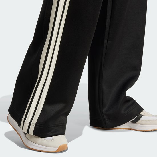 Voeten in zwarte trainingsbroek met wijde pijpen en beige 3-strepen, met sneakers.