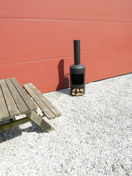 Kleine zwarte buitenhaard met houtblokken, op een grind ondergrond naast een rode muur en houten bank.