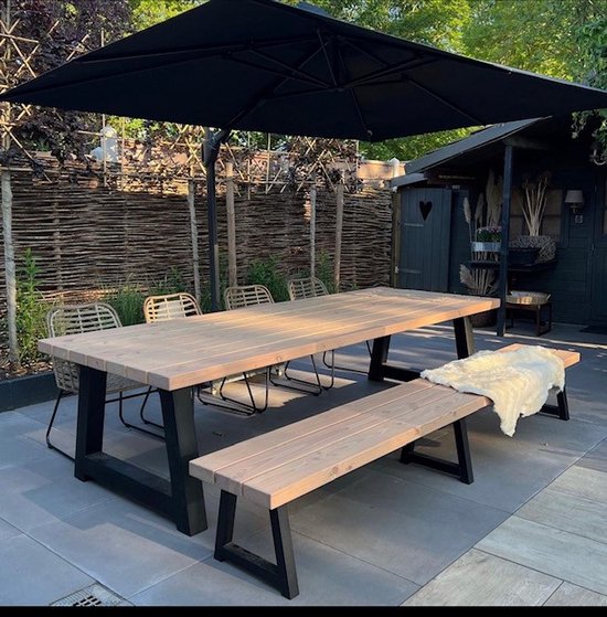 Houten tuintafel met bankje in een tuin onder een zwarte parasol, met stoelen en een schapenvacht op de bank.