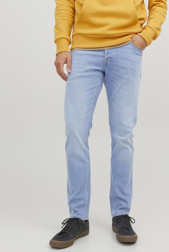Model draagt lichtblauwe slim fit jeans, gele hoodie en zwarte sneakers.