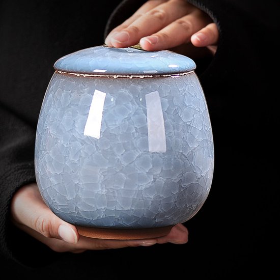 Handen houden een hemelsblauwe keramische pot met deksel vast. De pot heeft een krakelglazuur.