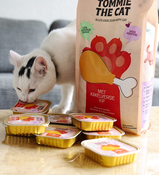 Witte kat met zwarte vlek bij het oor snuffelt aan blikjes Tommie the Cat kittenvoer naast een grote zak.