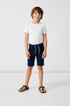 Jongen in donkerblauwe korte broek met rolzomen, wit t-shirt en sandalen.