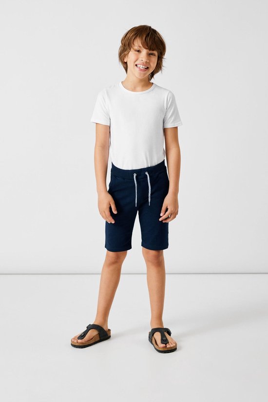 Jongen in donkerblauwe korte broek met rolzomen, wit t-shirt en sandalen.