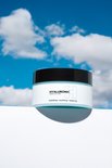 Glazen pot met lichtblauw hyaluronzuur masker op een witte kubus onder een blauwe hemel met wolken.