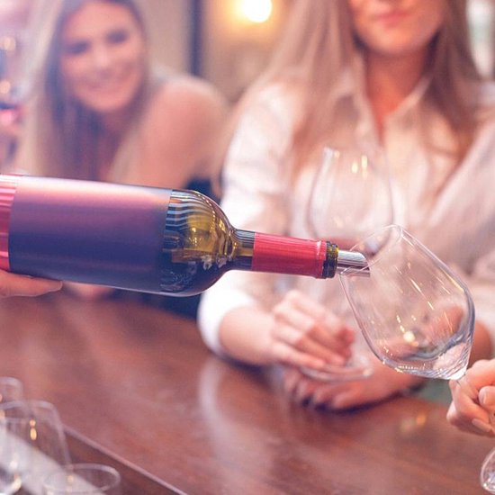 Een hand schenkt rode wijn uit een fles in een glas, twee vrouwen kijken toe.