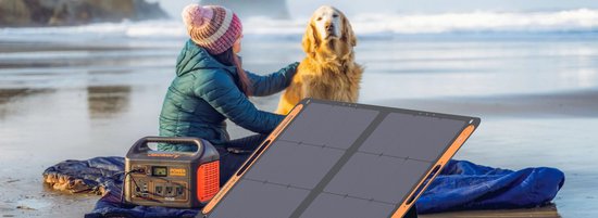 Op het strand ligt een Jackery zonnepaneel naast een power station. Een vrouw met een muts aait een hond.