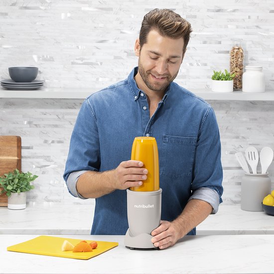 Man in blauw denim shirt maakt een oranje smoothie in een linnen beige Nutribullet.