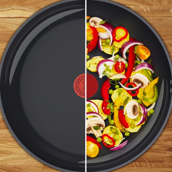 Een zwarte koekenpan, gevuld met verse groenten zoals aardappelen, paprika, ui, champignons en tomaatjes. De helft van de pan is leeg.