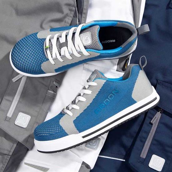 Twee Ardon werkschoenen, één blauw en grijs, liggend op werkkleding.