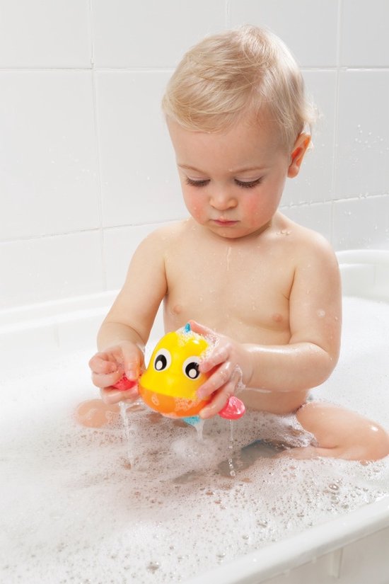 Een baby met blond haar in bad speelt met een gele badeend visje met rode vinnen.