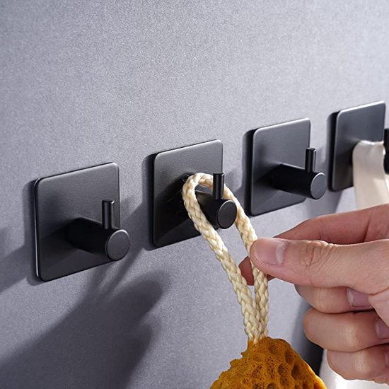 Vier zwarte haken aan een grijze muur, een hand wordt gebruikt om een badspons op te hangen.