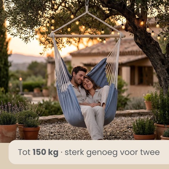 Een man en vrouw relaxen in een blauwgrijze hangstoel, opgehangen aan een boom met kerstverlichting.
