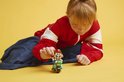 Een kind met een rode jas bouwt aan een groene Lego-motor met Mario erop.