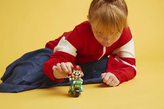 Een kind met een rode jas bouwt aan een groene Lego-motor met Mario erop.