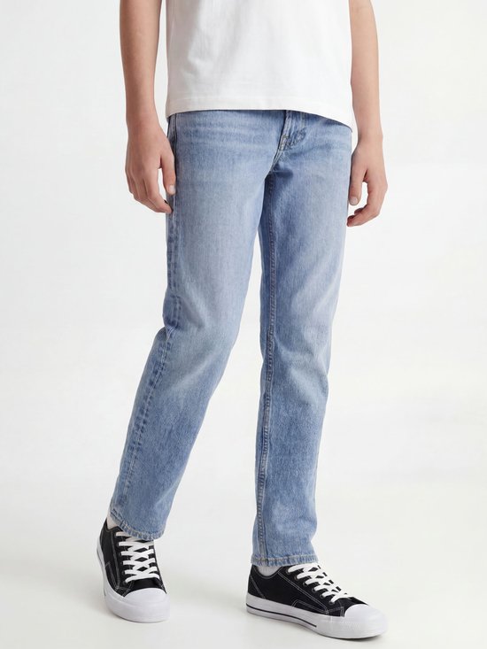 Jongen in wit T-shirt en lichtblauwe jeans, zittend. Draagt zwarte sneakers met witte zolen.