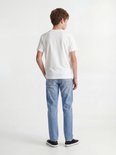 Jongen in wit T-shirt en lichtblauwe jeans, staand met handen in zakken. Draagt sneakers.