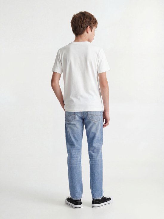 Jongen in wit T-shirt en lichtblauwe jeans, staand met handen in zakken. Draagt sneakers.