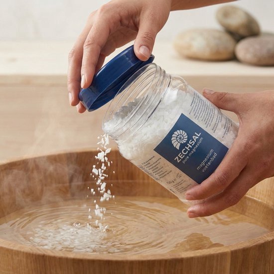 Een hand giet magnesiumvlokken uit een pot in een houten voetenbad met warm water.